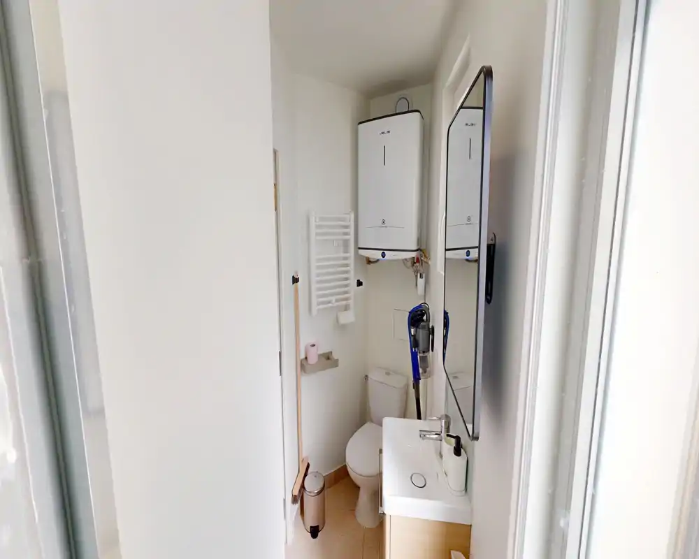 Salle de bains compacte et lumineuse avec toilettes, petit lavabo avec meuble, chauffe-eau mural, miroir et porte-serviettes chauffant ; propre et fonctionnelle.