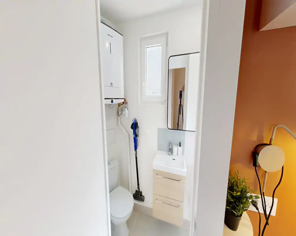 Petite salle de bains moderne avec toilettes, petit meuble vasque, miroir et chaudière murale ; espace lumineux et bien éclairé, idéal pour un petit appartement.