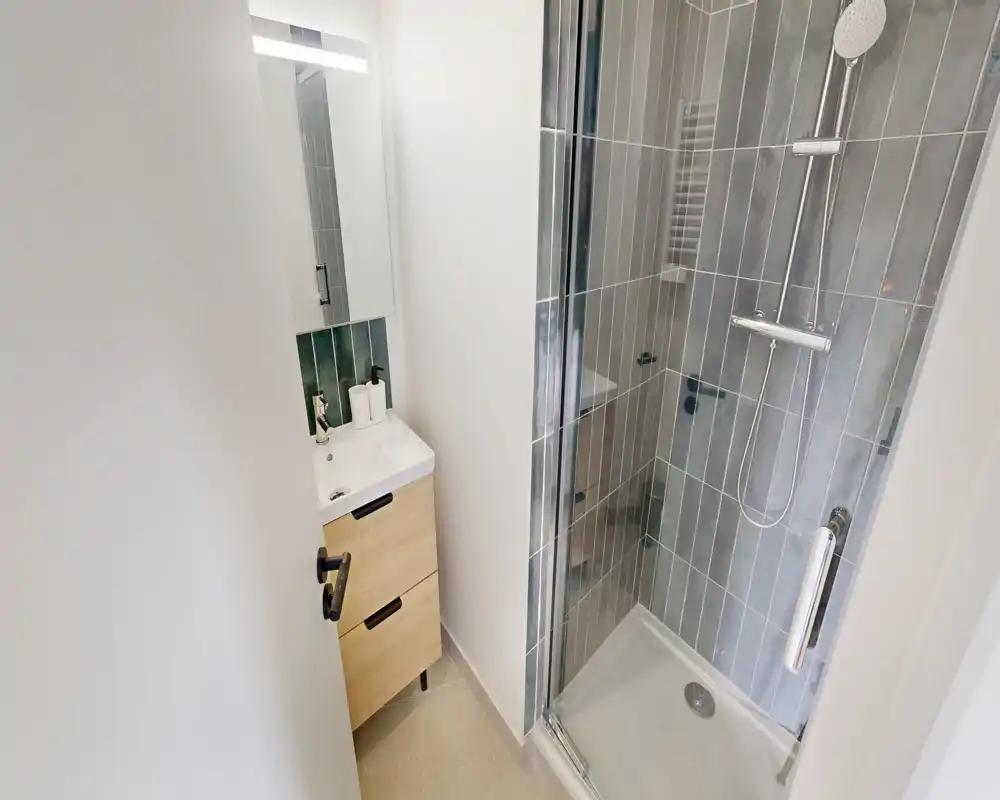Salle de bains moderne et compacte avec douche carrelée, porte en verre et petit meuble-vasque avec miroir — claire, propre et fonctionnelle.