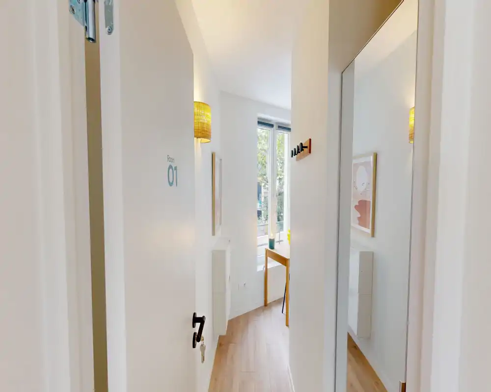 Entrée étroite et lumineuse avec une porte blanche, des patères murales et une grande glace reflétant le couloir ; sol en bois clair et un petit bureau visible au fond.