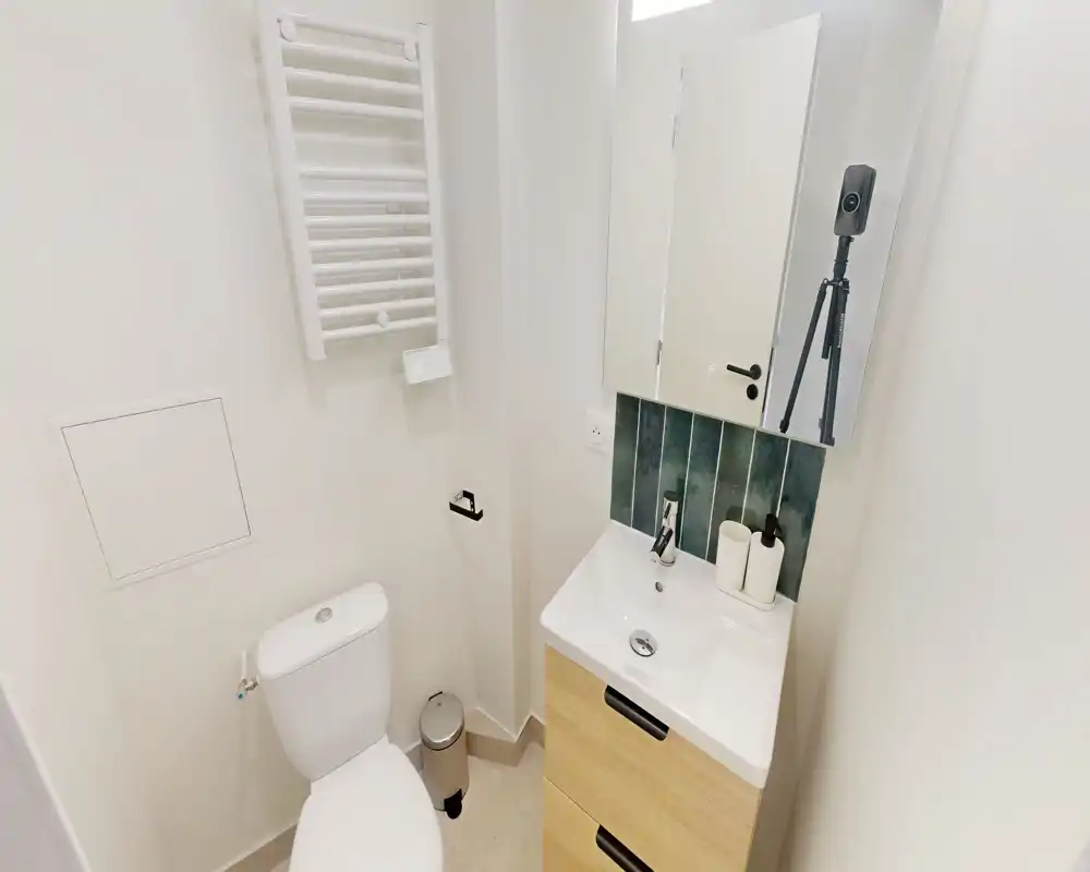 Petite salle de bains lumineuse avec WC, meuble vasque avec miroir, sèche-serviettes et rangement discret — finitions modernes et nettes, adaptée comme WC invité.