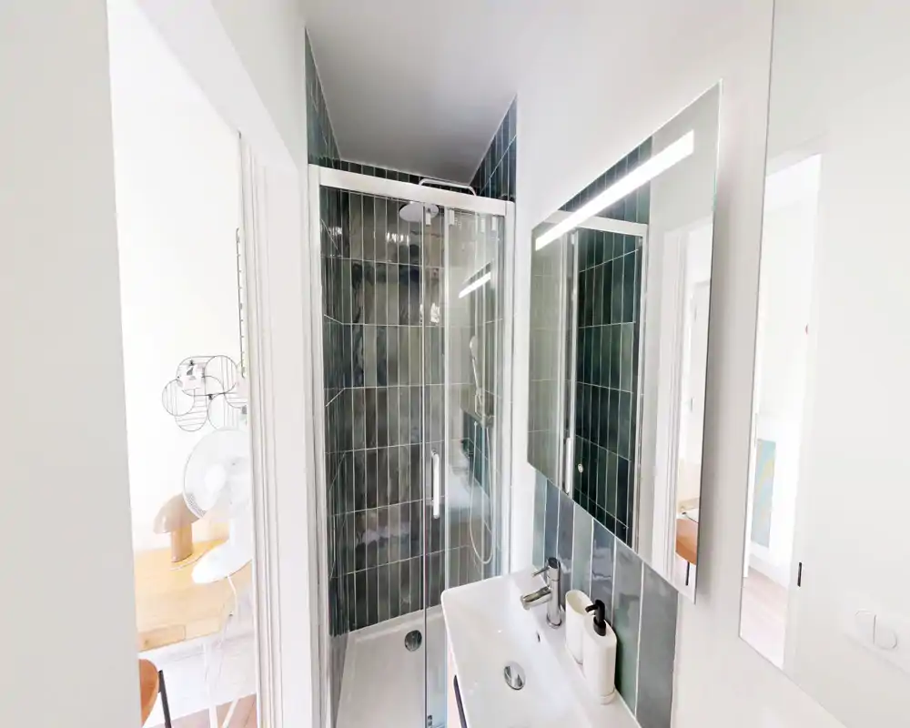 Salle de bains moderne et compacte avec cabine de douche vitrée, carrelage mural et petit lavabo avec miroir et distributeur—lumineuse, propre et bien agencée.