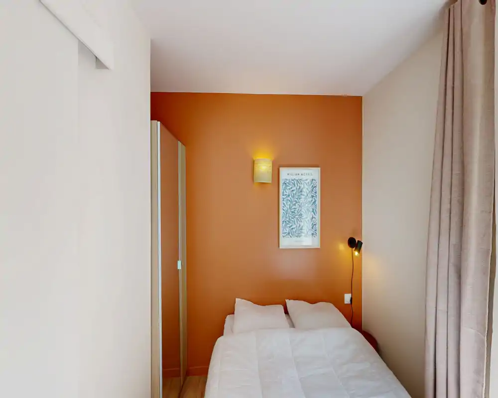 Chambre compacte et stylée avec un lit double devant un mur d'accent terracotta, lampes de chevet et une armoire mince—lumineuse et accueillante.
