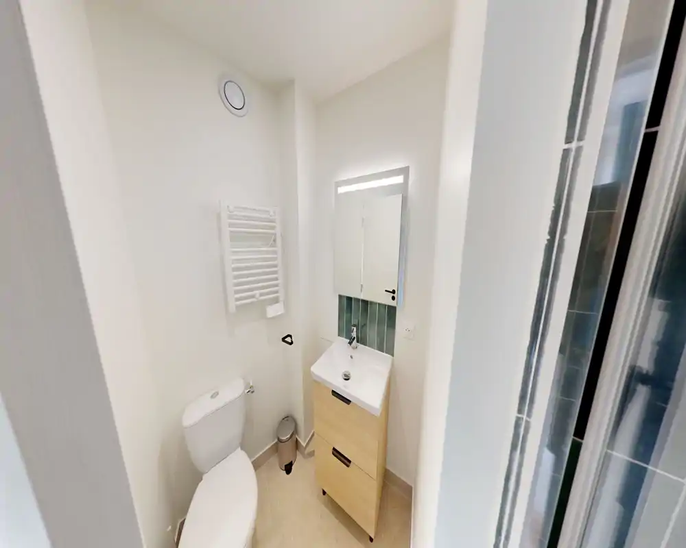 Salle de bain compacte et moderne avec WC, petit meuble vasque et miroir éclairé. Carrelage neutre et porte de douche en verre visible à droite.
