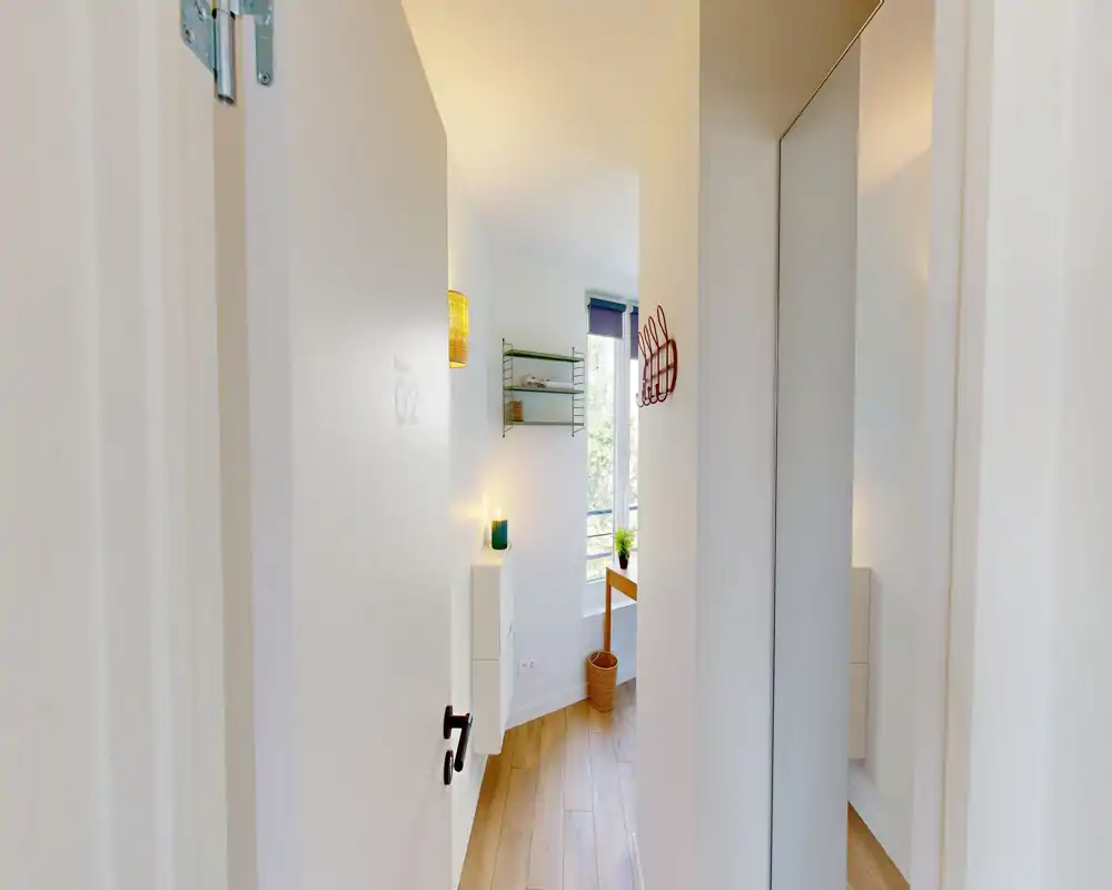 Entrée/couloir lumineux et étroit avec sol en bois, patères murales et miroir pleine hauteur. La lumière naturelle venant de la pièce adjacente donne une première impression accueillante.