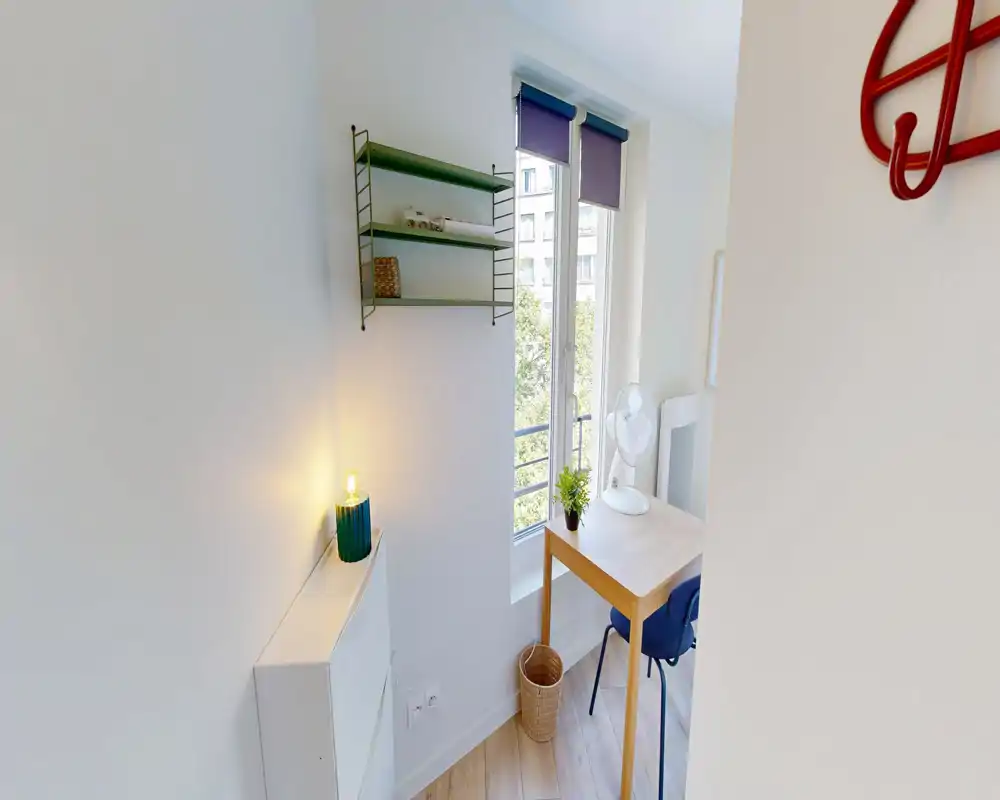 Coin lumineux et minimal du séjour avec un petit bureau en bois près de la fenêtre, chaise, ventilateur et étagère décorative ; belle lumière naturelle et style simple et soigné.