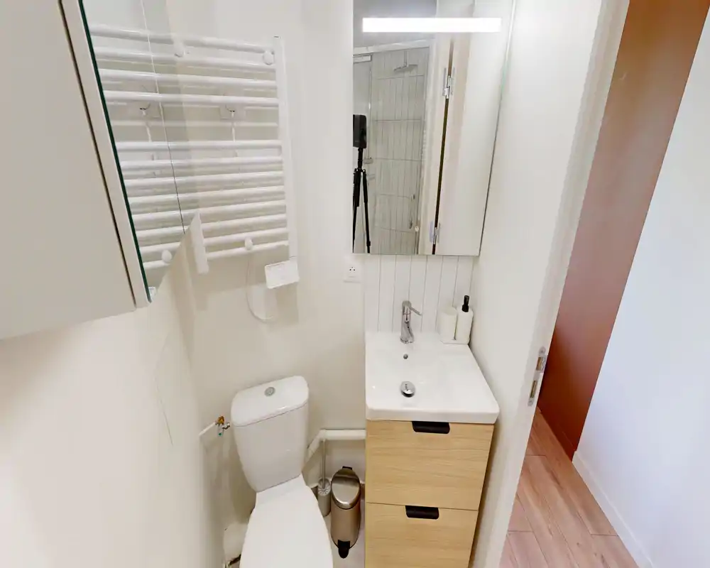 Salle de bain compacte et moderne avec WC blanc, petit meuble vasque à tiroirs en bois, miroir éclairé et radiateur porte-serviettes. Propre, bien éclairée et fonctionnelle.