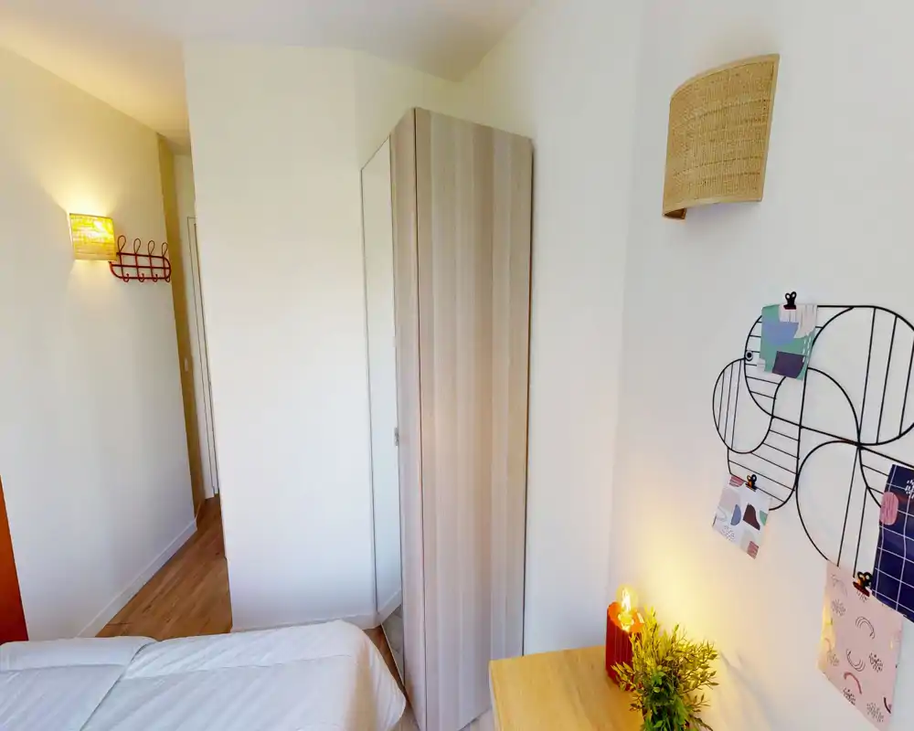 Coin chambre compact avec lit simple, grande armoire miroir et petit bureau en bois, grille murale décorative et éclairage chaleureux.
