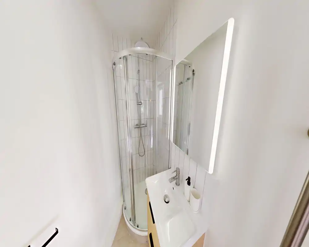 Salle de bains compacte et moderne avec cabine de douche en verre incurvée, miroir mural éclairé et petit lavabo avec robinetterie et accessoires.