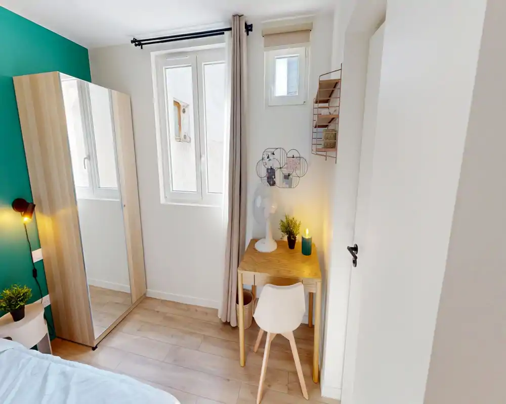 Chambre compacte et lumineuse avec un lit confortable, une armoire miroir et un petit bureau en bois près de la fenêtre — style moderne et épuré avec un mur vert et une belle lumière naturelle.