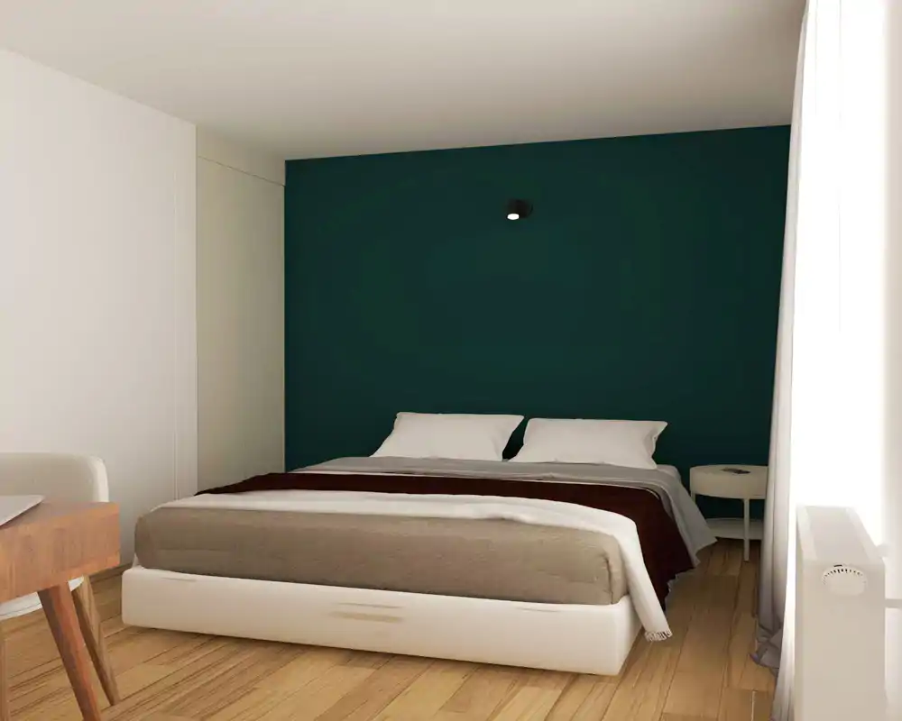 Chambre compacte et stylée avec un lit double centré devant un mur vert profond, oreillers moelleux et lumière naturelle venant de la fenêtre.