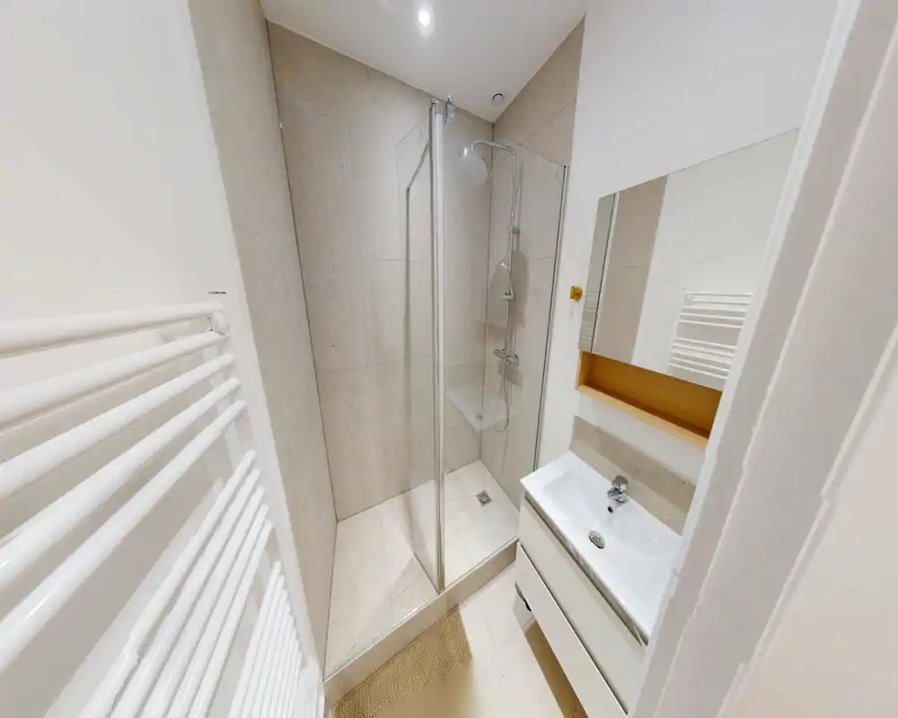 Bagno moderno e compatto con box doccia in vetro, lavabo a parete e piastrelle in pietra chiara — luminoso e pulito.