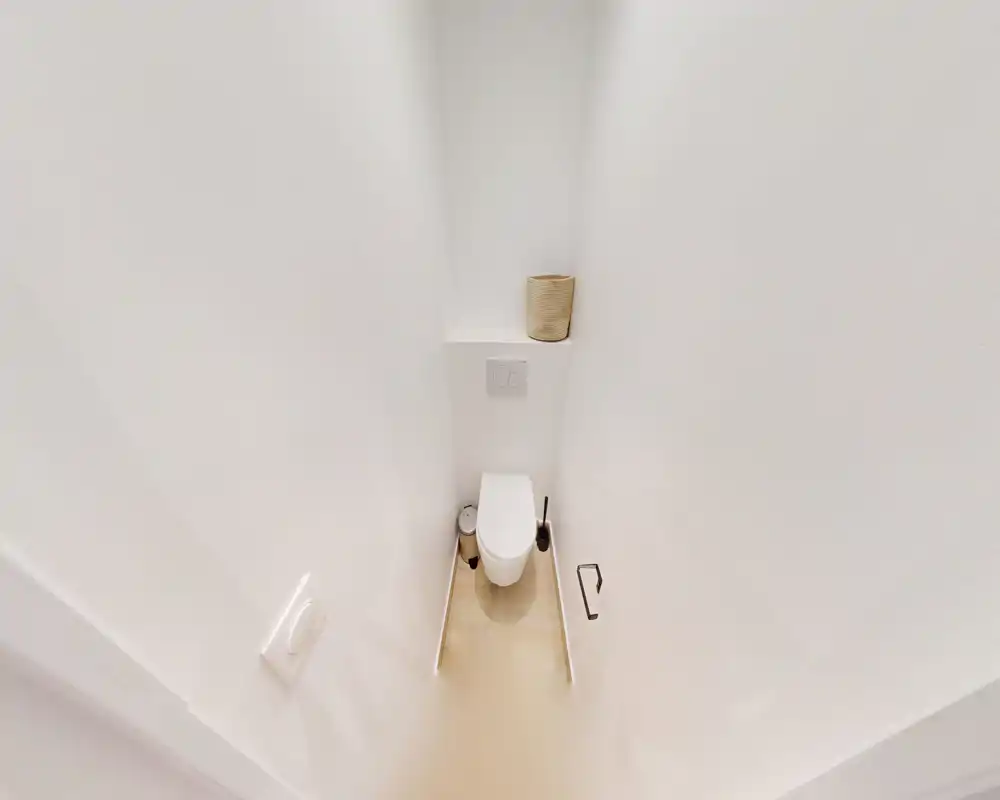 Bagno WC compatto e moderno con wc sospeso, placca di scarico incassata e finiture bianche neutre; un piccolo ripiano sopra la cassetta ospita un cesto decorativo.