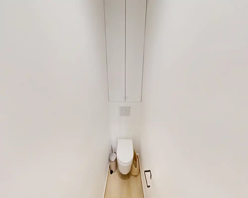 Piccolo bagno con WC, placca di scarico a parete, mobile sottile sopra e pavimento in legno chiaro. Stile minimal pulito, adatto come toilette per ospiti.