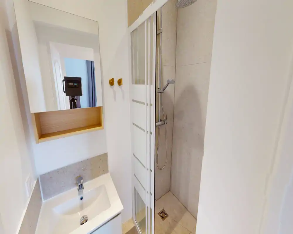 Salle de bains compacte et moderne avec douche à l'italienne, lavabo blanc et armoire-miroir — propre, lumineuse et bien entretenue.