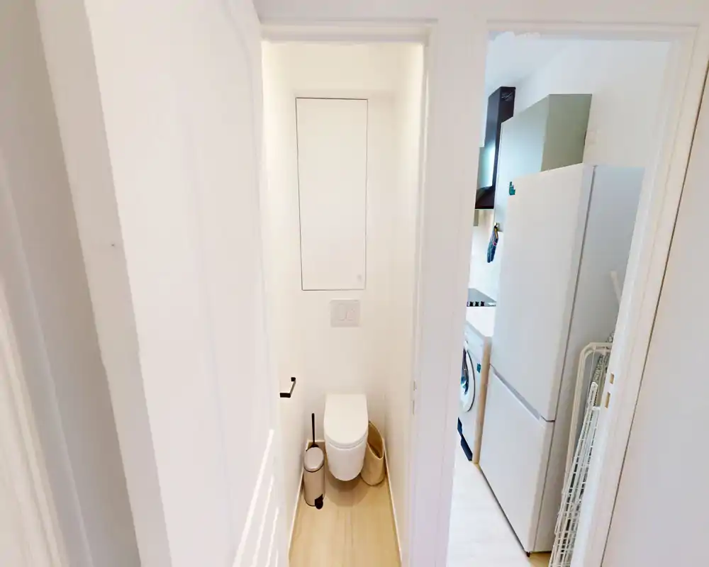 Toilettes séparées et compactes avec plaque de chasse d'eau murale, sol en bois clair et accessoires minimaux ; une zone buanderie/cuisine est visible par la porte.
