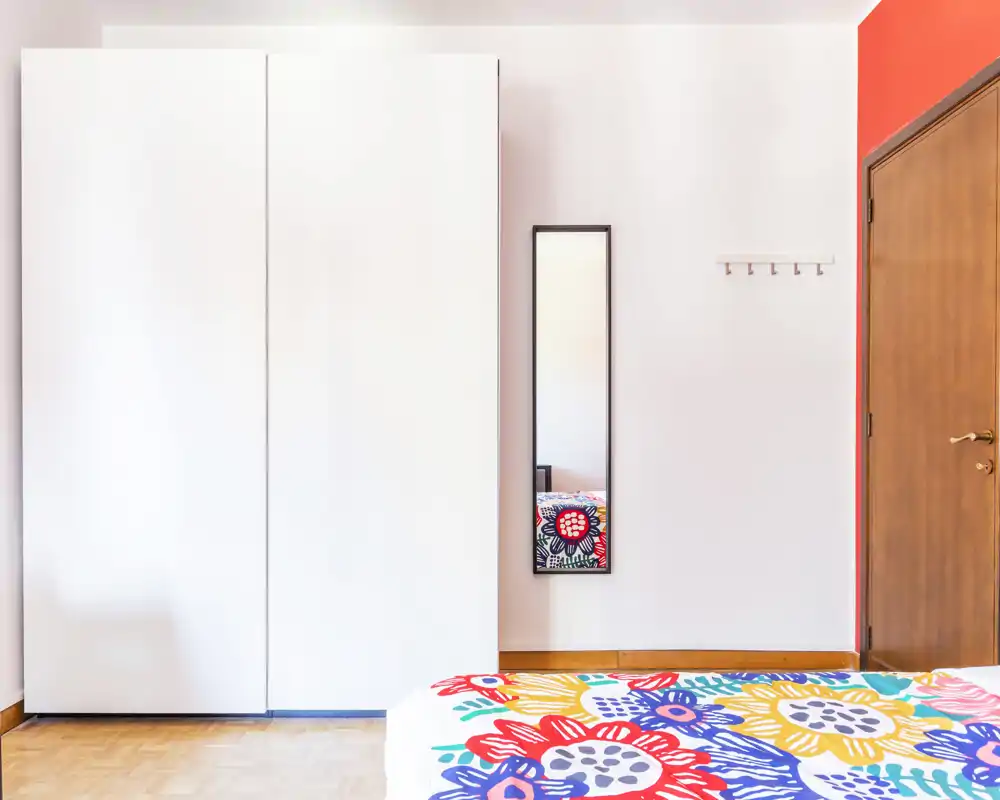 Camera da letto luminosa e moderna con copriletto colorato a fantasia, grande armadio bianco e specchio a figura intera — stile pulito e pavimento in parquet caldo.