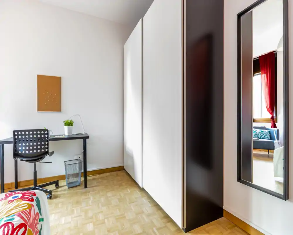 Angolo camera luminoso e moderno con scrivania, ampia armadiatura bianca e letto parzialmente visibile con biancheria colorata. Luce naturale e pavimento in parquet pulito rendono la stanza accogliente per affitti brevi.