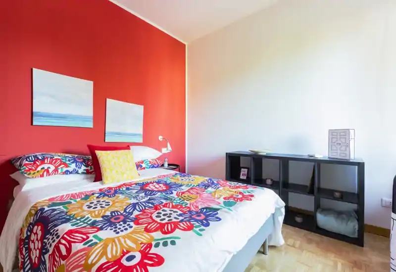 Camera da letto luminosa e colorata con un letto matrimoniale con biancheria floreale vivace su una parete d'accento rossa; lampada da comodino e scaffalatura bassa completano l'ambiente accogliente.