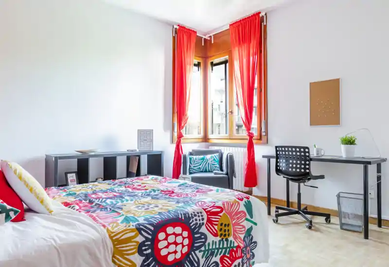 Camera da letto luminosa e colorata con un ampio letto coperto da una trapunta floreale, una scrivania e sedia vicino alla finestra e una poltrona accogliente — perfetta per un annuncio di affitto vivace e ben illuminato.