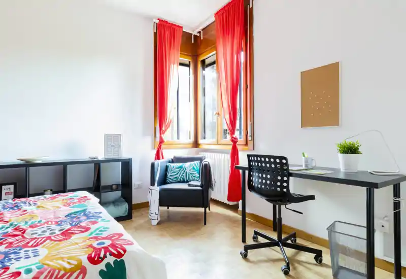 Camera da letto luminosa e colorata con copriletto decorato, una comoda poltrona vicino alla finestra e scrivania ordinata—ben illuminata e allestita per annunci.