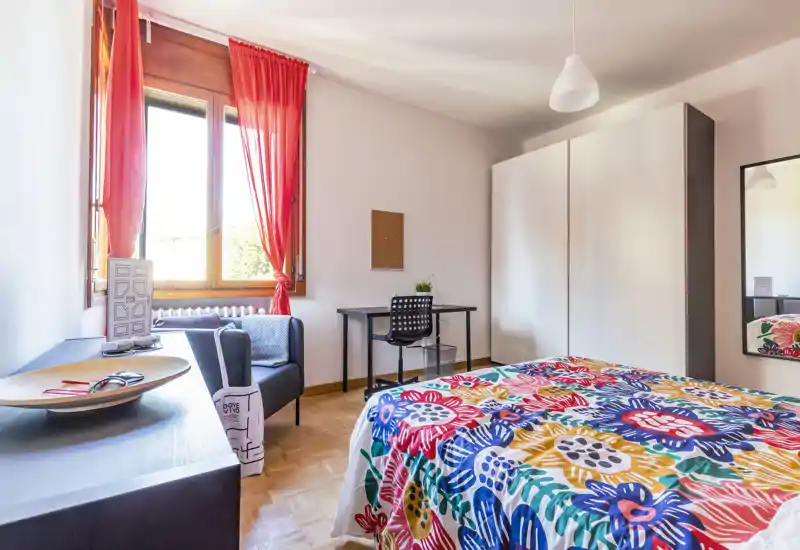Camera da letto luminosa e ben arredata con una coperta floreale colorata, armadio, scrivania e una poltrona accogliente vicino alla finestra che lascia entrare molta luce naturale.