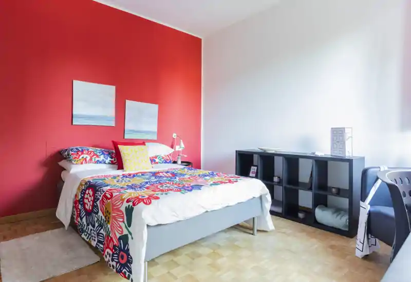 Camera da letto luminosa e colorata con letto completo, biancheria decorata e una parete rossa a contrasto; mensole ordinate e luce naturale la rendono accogliente.