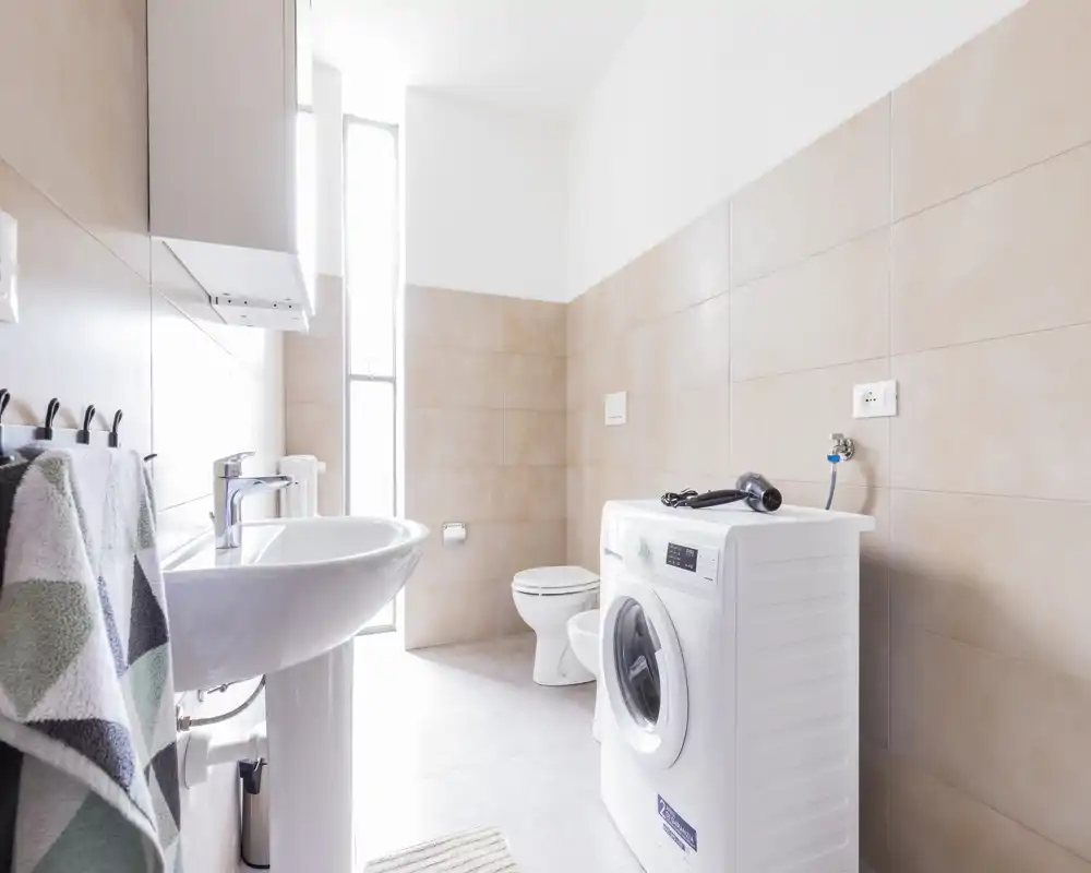 Bagno moderno e luminoso con lavabo a colonna, WC e lavatrice a carica frontale. Piastrelle beige neutre e una finestra alta garantiscono molta luce naturale.