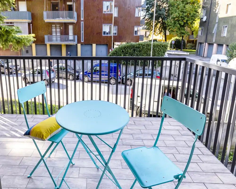 Piccolo balcone soleggiato con tavolino bistro in metallo turchese e due sedie abbinate; pavimentazione in piastrelle e affaccio sulla strada, ideale per il caffè mattutino.