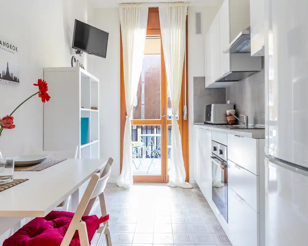 Cucinotto luminoso e moderno con mobili bianchi, forno incassato, piccolo tavolo da pranzo e porta sul balcone che fa entrare molta luce naturale — spazio ordinato ideale per affitti brevi.
