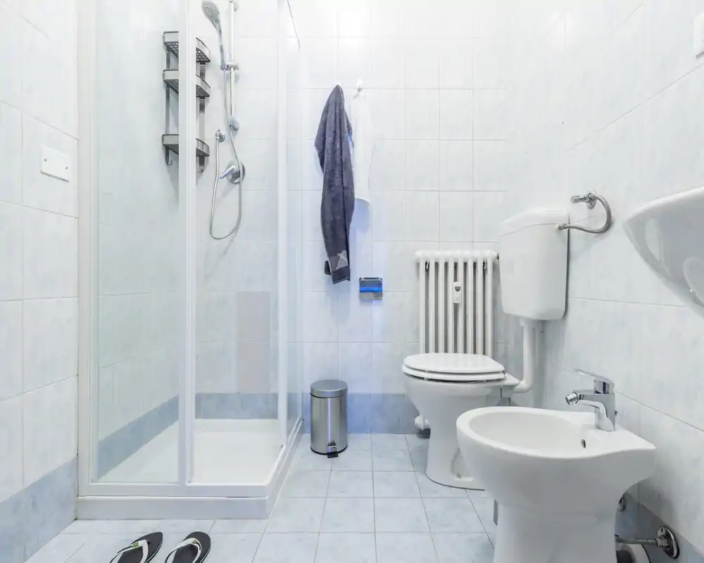 Bagno pulito e luminoso con box doccia, WC, bidet e lavabo in piastrelle azzurre. Asciugamani appesi e un piccolo cestino vicino alla doccia.