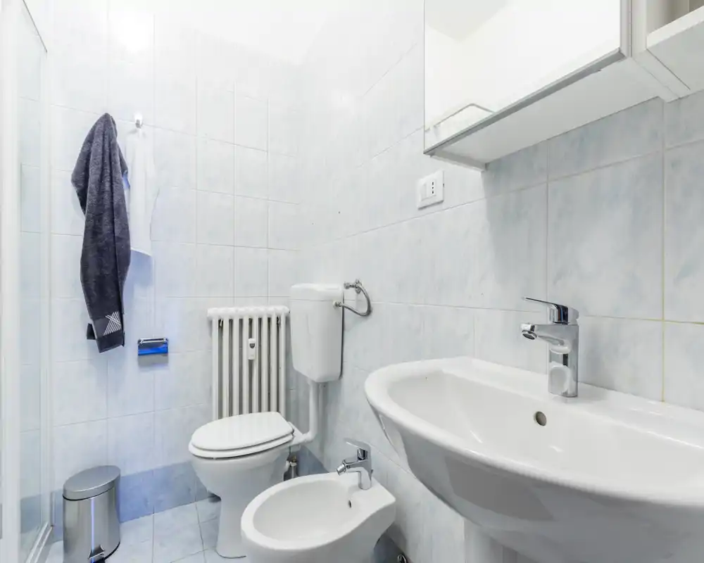 Bagno compatto e pulito con lavabo, bidet e wc su piastrelle azzurro chiaro; illuminazione luminosa e rubinetteria cromata moderna.
