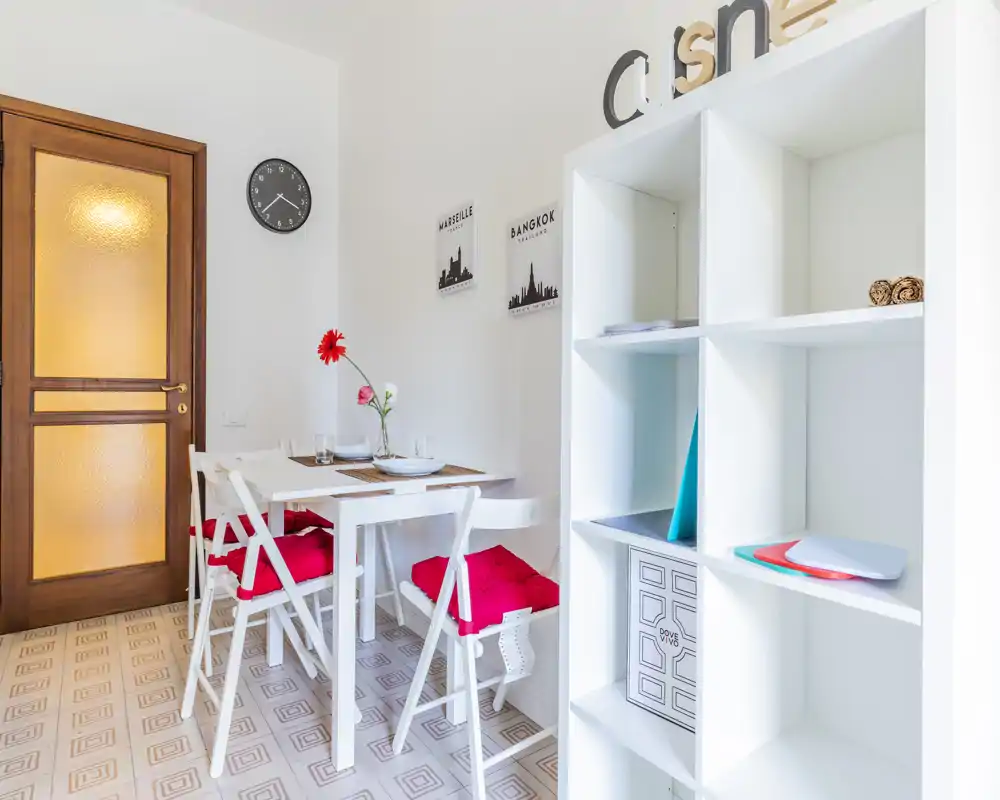 Angolo pranzo compatto e curato con tavolo bianco e quattro sedie pieghevoli con cuscini rossi, quadri decorativi e un fiore fresco al centro — luminoso e accogliente per pasti informali.