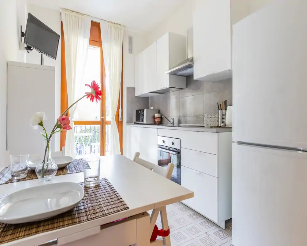 Cucina angolare moderna e luminosa con mobili bianchi, forno incassato e un piccolo tavolo apparecchiato per due; la luce naturale dalla porta sul balcone rende l'ambiente arioso e accogliente.