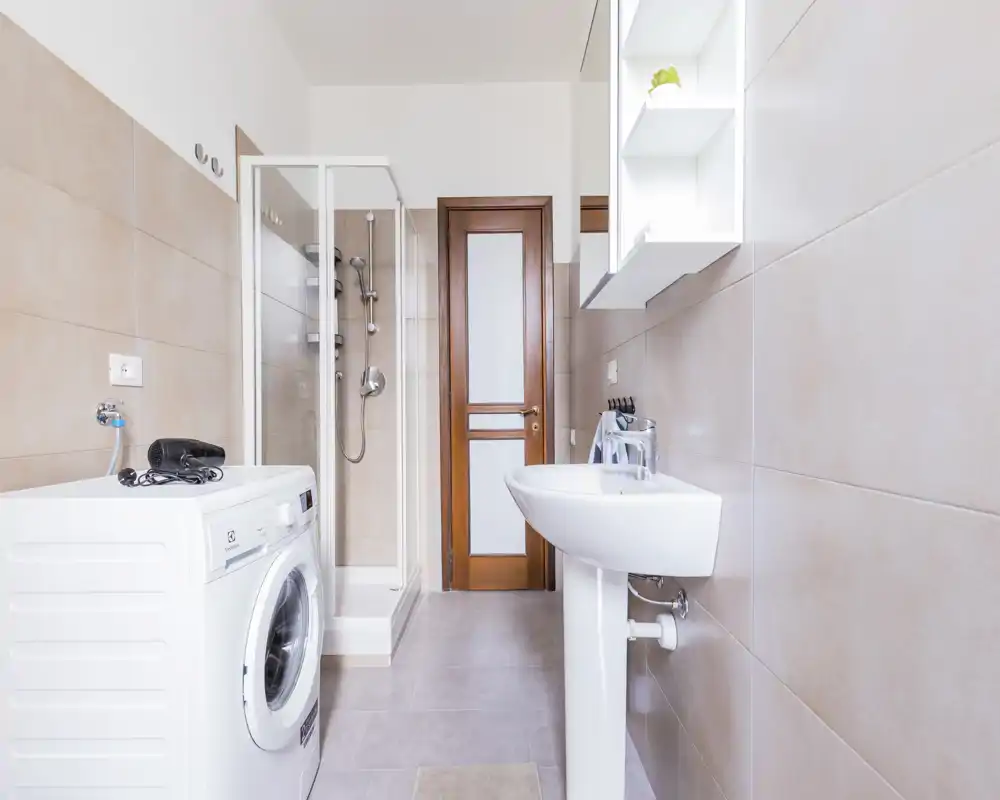 Bagno moderno e luminoso con cabina doccia, lavabo a colonna e lavatrice. Piastrelle dai toni neutri e illuminazione chiara creano un ambiente pulito e arioso.