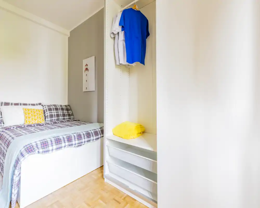 Dormitorio luminoso y bien presentado con cama forrada de cuadros, cojín decorativo y armario abierto; suelo de parquet y paredes neutras que aportan una sensación fresca y acogedora.