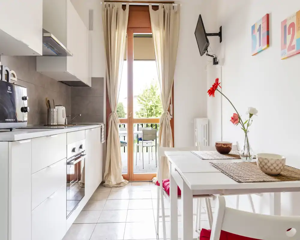 Cocina moderna y luminosa con armarios blancos, horno empotrado, pequeños electrodomésticos en la encimera y una mesa junto a la puerta balcón que aporta mucha luz natural.