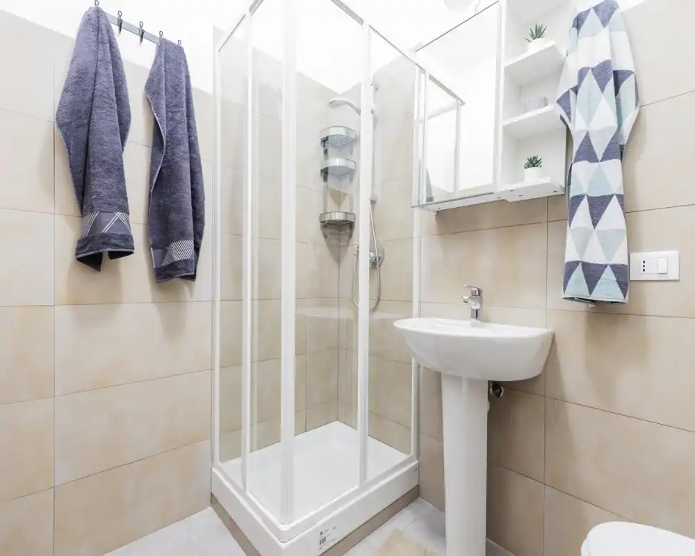 Baño moderno y luminoso con ducha de esquina acristalada, lavabo con pedestal y percheros para toallas: espacio limpio y bien iluminado ideal para el anuncio.