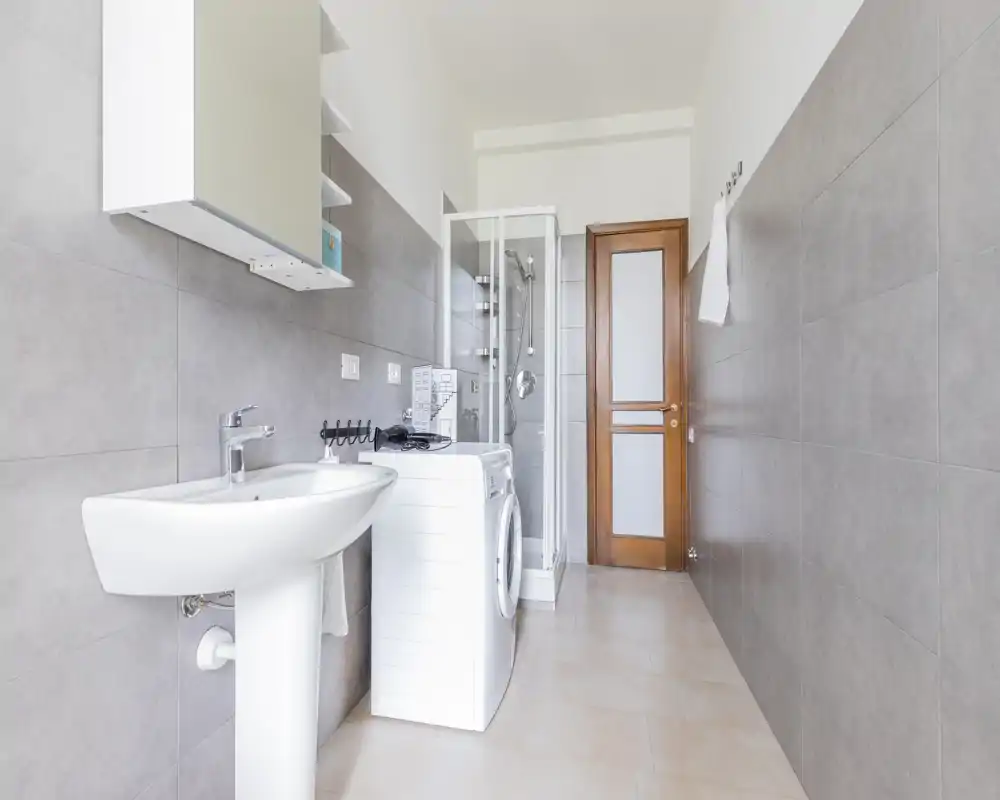 Baño moderno y luminoso con lavabo, cabina de ducha y lavadora; azulejos gris claro y puerta de madera que crean un espacio funcional y limpio.