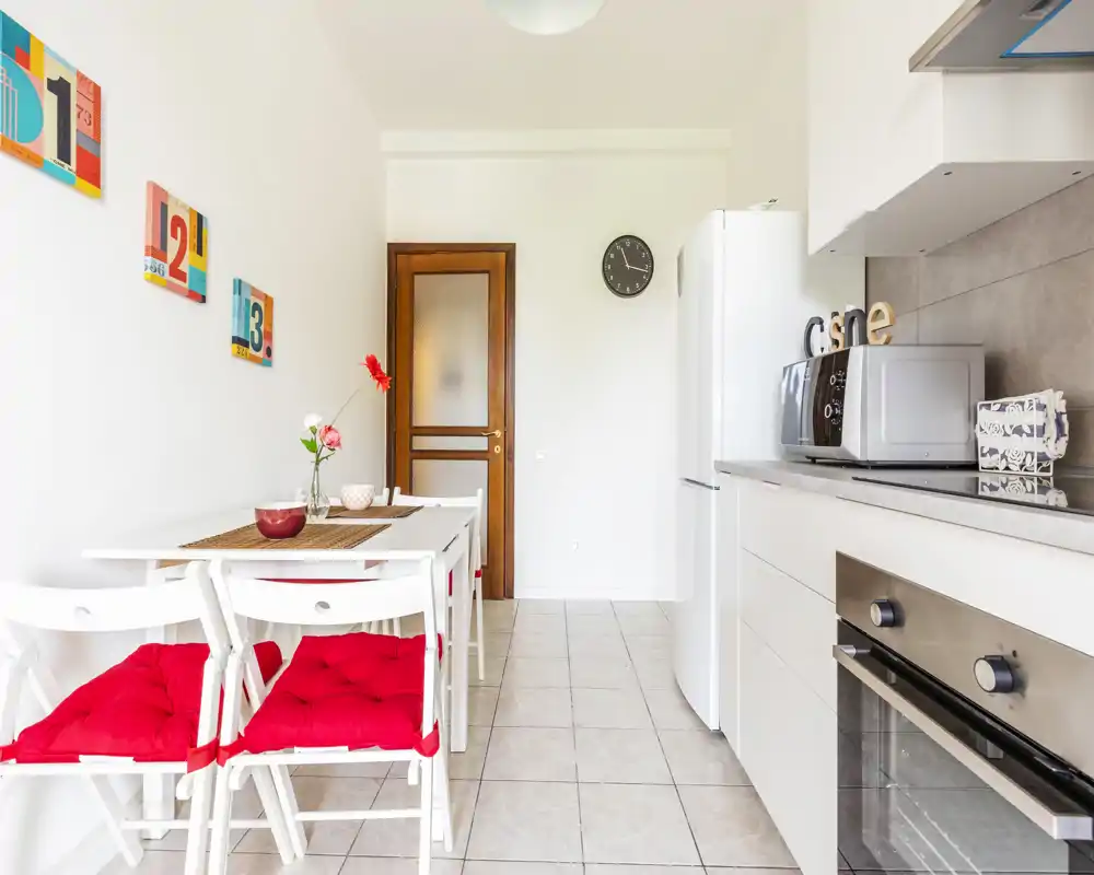 Cocina moderna y luminosa con un rincón para comer compacto: muebles blancos, horno empotrado, nevera y mesa pequeña con cojines rojos. Estética limpia y aireada, ideal para alquileres urbanos.