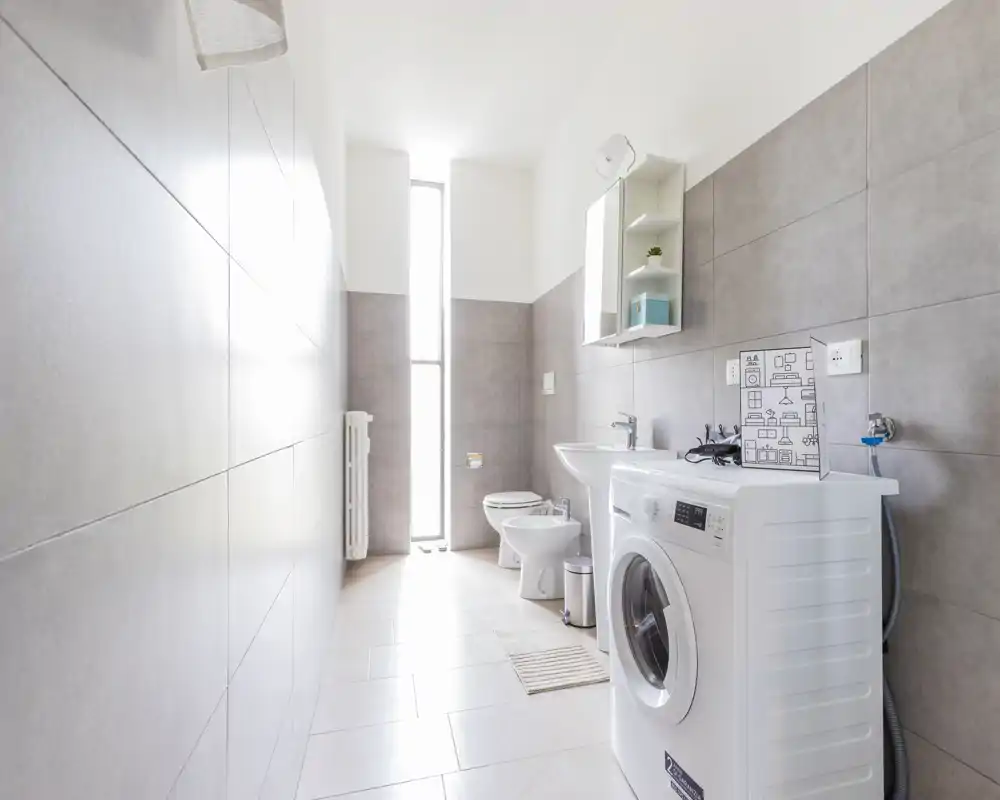 Baño moderno y luminoso con lavadora, lavabo, bidé y WC. Azulejos grises y una ventana estrecha y alta proporcionan abundante luz natural.