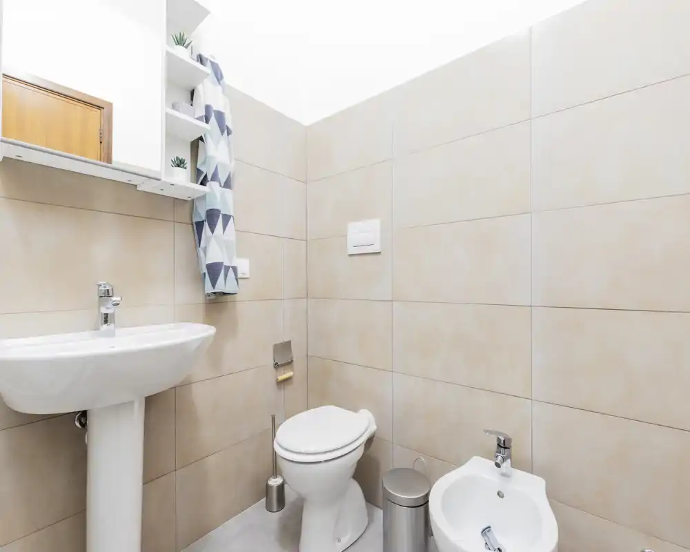 Baño moderno y luminoso con lavabo de columna, inodoro y bidé; azulejos beige neutro y grifería cromada ofrecen un aspecto limpio y minimalista.