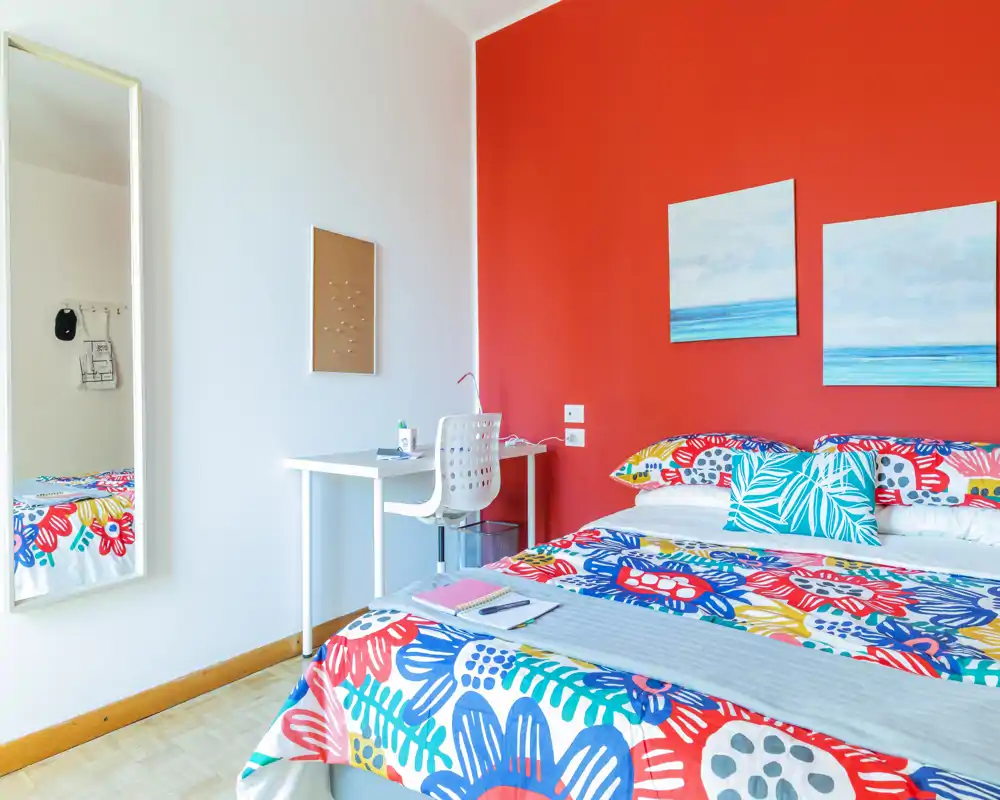Camera da letto luminosa e colorata con letto matrimoniale con biancheria floreale vivace, una piccola scrivania bianca e uno specchio a tutta parete. La parete rossa e i quadri coordinati danno un aspetto moderno e accogliente perfetto per annunci.