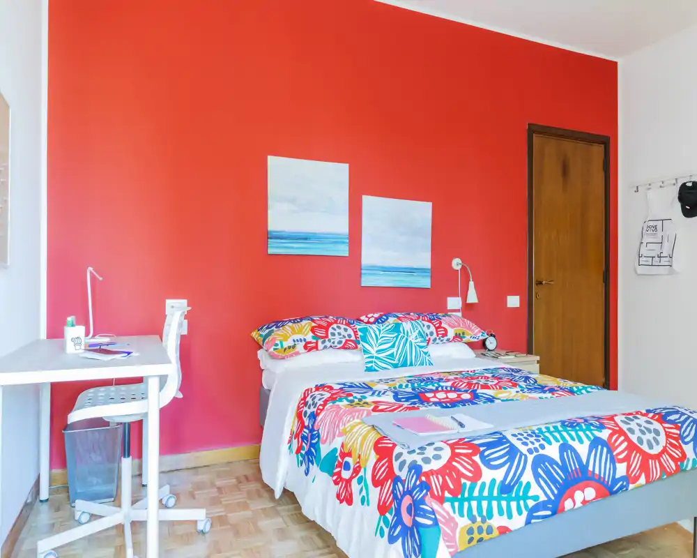 Camera da letto luminosa e vivace con letto matrimoniale con biancheria floreale colorata, lampada da comodino e quadri su una parete accentuata rossa; a sinistra è presente una scrivania con sedia per lavorare.