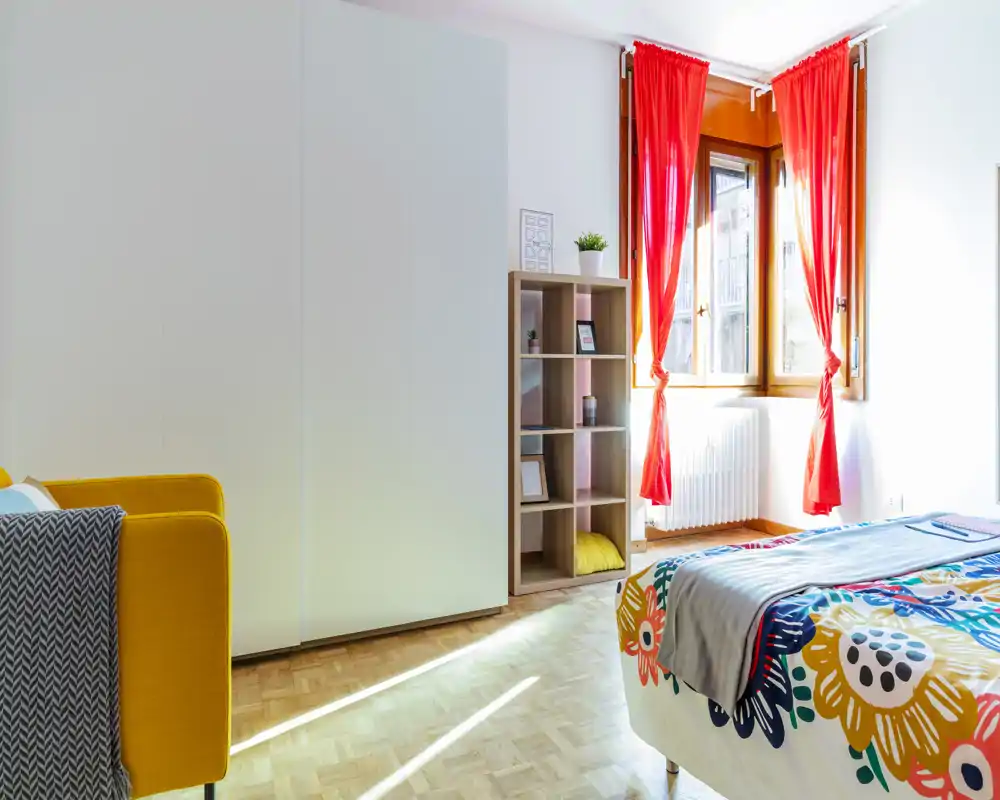 Camera da letto luminosa e colorata con piumone fantasia, grande finestra con tende rosse, armadio e scaffale; ambiente moderno, soleggiato e accogliente, ideale per annunci.