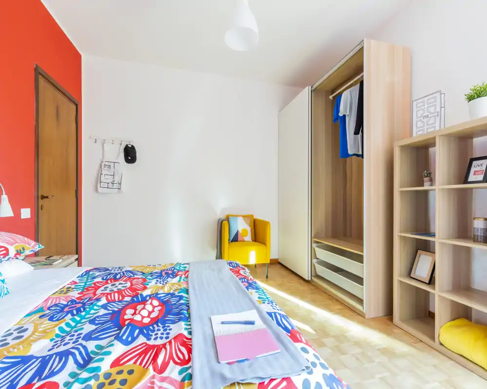 Camera da letto luminosa e colorata con grande copriletto a fantasia, armadio aperto e una poltrona gialla accogliente — ben illuminata e arredata con gusto.
