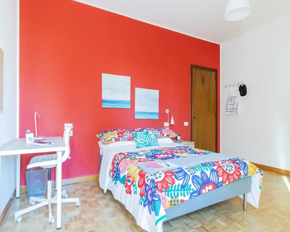 Camera da letto luminosa e colorata con letto matrimoniale, copriletto floreale vivace, scrivania e lampada da comodino su una parete rossa accesa.