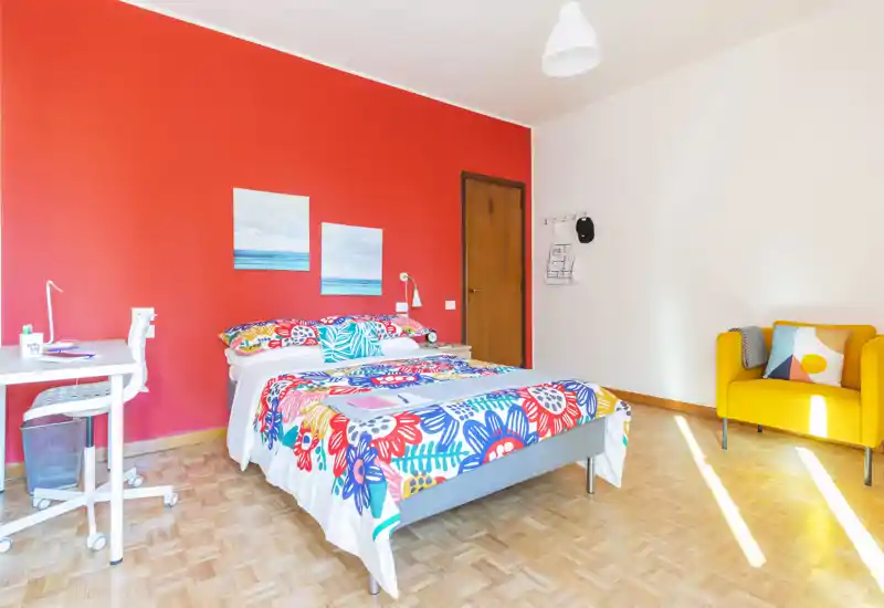 Camera da letto luminosa e colorata con letto completo, biancheria vivace, angolo studio e una poltrona gialla accogliente. La luce naturale e la parete a colori rendono l'ambiente invitante per gli annunci.