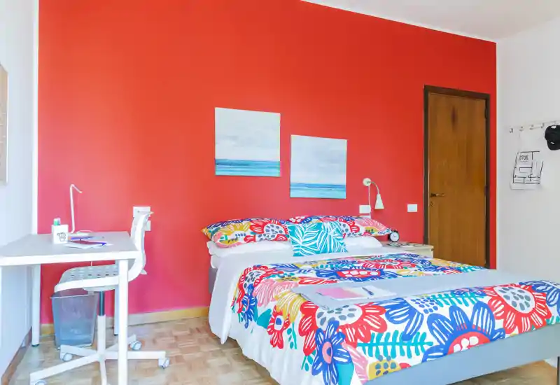 Camera da letto luminosa e vivace con letto matrimoniale con biancheria floreale colorata, lampada da comodino e quadri su una parete accentuata rossa; a sinistra è presente una scrivania con sedia per lavorare.