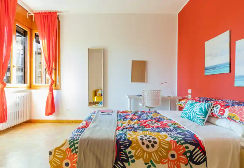 Camera da letto luminosa e colorata con letto matrimoniale con copripiumino floreale, scrivania e sedia contro una parete corallo e grandi finestre che lasciano entrare molta luce naturale.